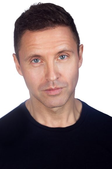 Man with short dark hair, wearing a black shirt, facing the camera with a neutral expression.