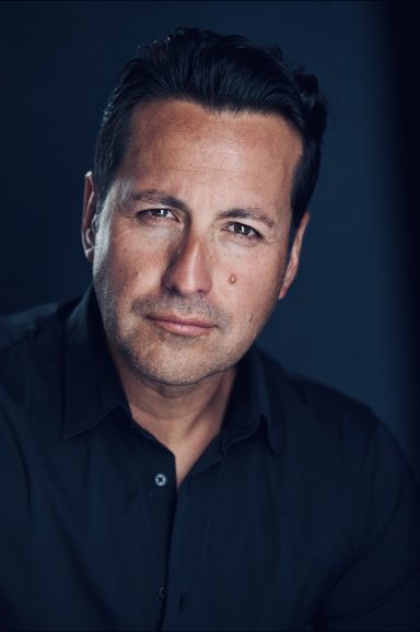 Man with short dark hair, serious expression, wearing a black shirt against a dark background.