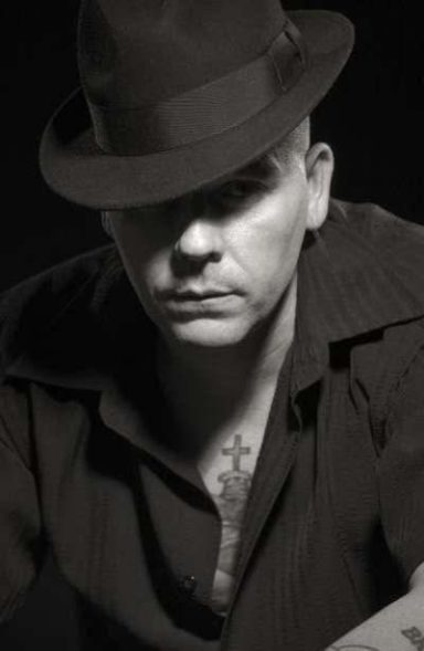 Man in a black shirt and hat, with a serious expression and visible tattoo on his chest.