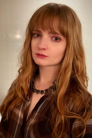 A young woman with long, wavy hair and a plaid shirt poses, looking thoughtfully at the camera.