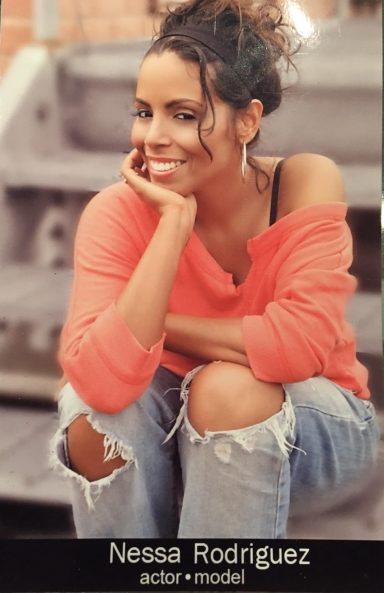 Smiling woman in an orange sweater sitting on stairs, with curly hair and casual jeans.