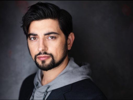 Headshot of a man with dark hair and a beard, wearing a hoodie and jacket, looking confidently.